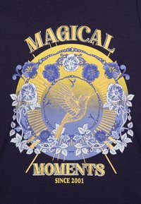 Mørk lilla t-shirt med et farverigt motiv af en fugl og blomster, med gule og blå accenter, samt teksten "MAGISKE ØJEBLIKKE SEDAN 2001."