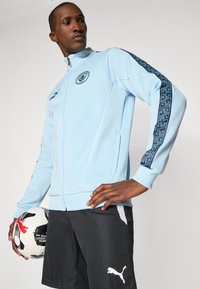 Light blue zip-up jacket with dark pattern accents on sleeves, white trims, logo on chest, paired with black shorts and a black-and-white soccer ball.