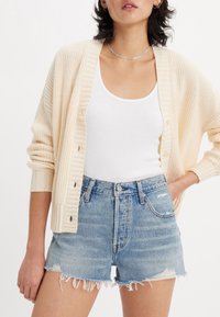 Cárdigan de punto beige claro con textura acanalada y botones, combinado con una camiseta sin mangas blanca y pantalones cortos de mezclilla desgastados en azul claro.