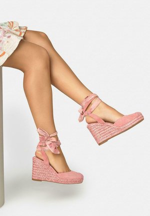 Piernas con sandalias cuña de esparto rosas con lazos en el tobillo, cruzadas y elevadas sobre un fondo liso.