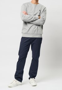 Hombre de pie con los brazos cruzados, vistiendo un sudadera gris, pantalones azul marino y zapatillas blancas, frente a un fondo blanco liso.