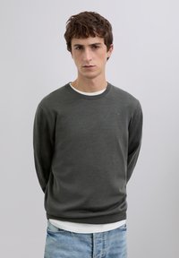 Dunkelgrauer Pullover mit rundem Ausschnitt und langen Ärmeln, verziert mit einem dezenten, eingeprägten Logo. Trägt man über einem weißen Hemd, kombiniert mit hellblauen Jeans.
