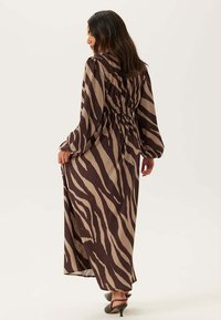 Maxi šaty v hnědém a béžovém vzoru zebřích pruhů, s dlouhými rukávy, elastickým pasem a splývavou látkou. Nošeny s hnědými lodičkami na podpatku.