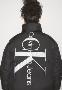 Giacca a collo alto in nylon nero, con grande logo bianco "Calvin Klein Jeans" sul retro in allineamento verticale e dettagli riflettenti.