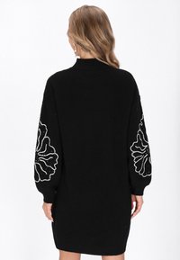 Schwarzes Strickkleid mit hohem Halsausschnitt, langen Ärmeln mit weißen gestickten Blumenmustern und lockerer Passform, das bis zur Mitte des Oberschenkels reicht.