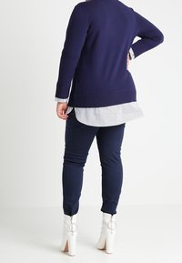 Marineblauer Strickpullover mit einem langen, gestreiften Hemdschnitt, kombiniert mit marineblauen Hosen und weißen Knöchelboots mit runden Hardware-Akzenten.