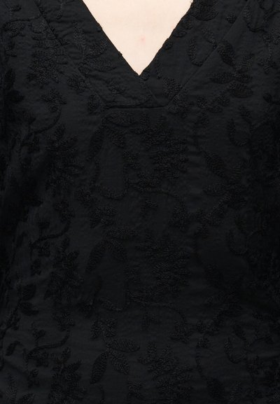 Haut en tissu noir avec un col en V et une broderie florale. Présente des motifs texturés dans un design ton sur ton, avec une apparence douce et légère.