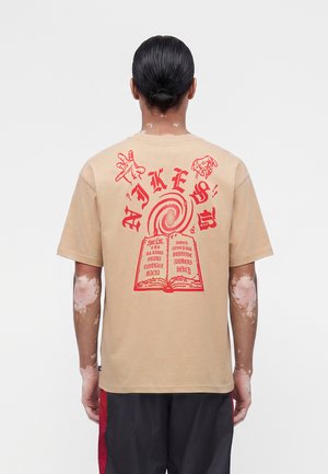 Personne aux cheveux en chignon portant un t-shirt beige avec un graphisme rouge représentant des mains, une spirale et un livre ouvert avec du texte dans le dos, debout devant un fond blanc.