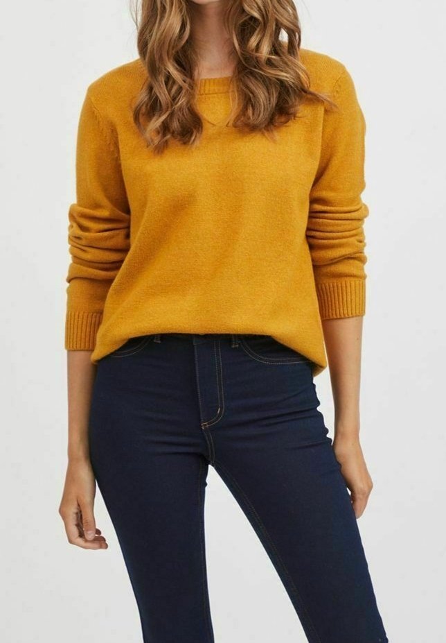 Senapsgul stickad tröja med rund halsringning och ribbade ärmslut, kombinerad med mörkblå tajta jeans. Slät textur, avslappnad passform.