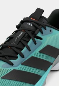 Τιρκουάζ αθλητικό παπούτσι με μαύρα κορδόνια, επάνω μέρος από δίχτυ, τρεις μαύρες ρίγες και πορτοκαλί λεπτομέρειες· διαθέτει υφή και διαπνέον σχεδιασμό.