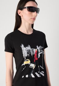 Must musta t-särk, millel on graafiline kujundus naisest punases taktis ja kõrgetes saapastes, ning kollane takso ja linnamaastik taustal.