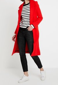 Manteau en laine rouge, chemise à rayures noires et blanches, jean noir et baskets blanches. Le manteau présente un col, des boutons et un design long.