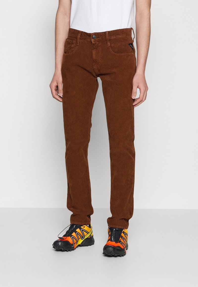 Personne portant un pantalon en velours côtelé marron, une chemise blanche et des chaussures de sport noires, orange et jaunes, debout sur un sol gris clair uni.