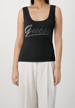 Vrouw draagt een mouwloze zwarte top met zilveren "Guess" logo en losse witte broek, staand tegen een effen achtergrond.