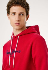 Rode hoodie van katoen, met een trekkoord met metalen punten en een marineblauw logo "Pepe Jeans LONDON" op de voorkant. Zachte textuur.