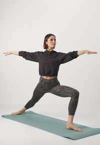 Felpa cropped nera con cappuccio, leggings grigi a fantasia e piedi nudi su un tappetino da yoga color teal. Modella in posizione del guerriero.