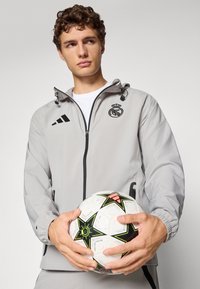 Lys grå vindjakke med sorte detaljer, der har hætte, lynlås og Real Madrid-logo. Holder en hvid fodbold med sort og grøn stjernedesign.