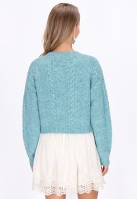 Suéter cortado de punto en azul claro con patrones de trenzado texturizados, escote redondeado y puños y dobladillo acanalados. Combinado con una falda blanca.