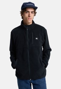 Giacca in pile nera con colletto alto, zip frontale e tasche laterali. Presenta un piccolo logo bianco sul petto e una texture morbida.