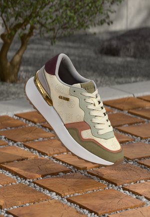 Cremefarbener und grüner Sneaker mit gemustertem Leder, Schnürdesign, goldenen Akzenten und einer strukturierten dicken Sohle, präsentiert auf braunen Fliesen und Kieselsteinen.