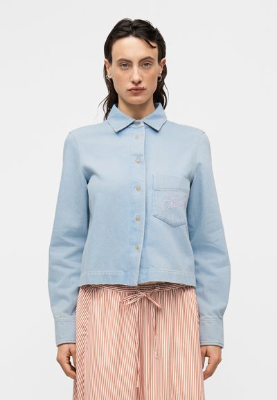 Light blue denim shirt with a pointed collar, front buttons, and a chest pocket featuring pink embroidered text; paired with striped skirt.