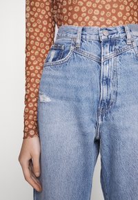 Vaqueros azul claro de talle alto con un parche desgastado, combinados con un top marrón de manga larga con un estampado floral. Textura de mezclilla visible.