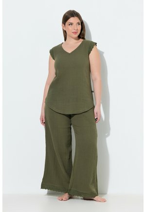 Femme portant un haut sans manches vert olive et un pantalon à jambes larges, debout pieds nus contre un mur blanc, regardant légèrement sur le côté.