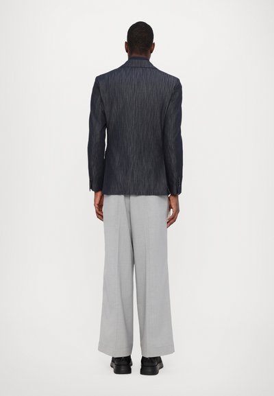 Navy-blau-gestreifter Blazer mit strukturierter Silhouette. Hellgraue, weit geschnittene Hose, kombiniert mit schwarzen Schuhen, vor einem schlichten weißen Hintergrund.