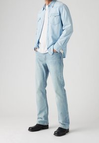 Chemise en denim bleu clair portée sur un t-shirt blanc, associée à un jean bleu clair et des chaussures noires. Design simple et coupe décontractée.