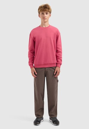 Tienerjongen die staat, met een roze sweatshirt, bruine broek en zwarte sneakers tegen een effen lichtgrijze achtergrond.