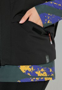 Schwarze Jacke mit einer Tasche mit Klettverschluss, ergänzt durch einen gemusterten Saum in Grün und Lila mit gelben Akzenten. Robustes Material.
