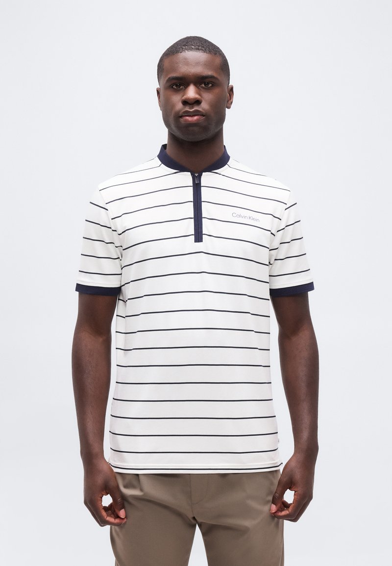 Άνδρας που φοράει λευκό κοντομάνικο πουκάμισο με λεπτές μαύρες οριζόντιες ρίγες, γιακά με φερμουάρ σε ναυτικό μπλε και χακί παντελόνι, στέκεται μπροστά σε μονόχρωμο φόντο.