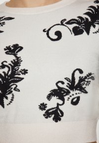 Maglione lavorato a maglia di colore crema con motivi floreali neri. Presenta un collo a girocollo a coste e una superficie liscia. Maniche di lunghezza media.