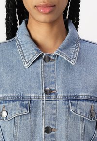 Lyseblå denimjakke med krage, synlige sømmer og fire metalldekknapper foran, med et klassisk design og tekstur.