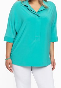 Top turchese, dalla vestibilità ampia, con colletto e maniche corte. Texture morbida, orlo dritto e dettagli delle cuciture discreti. Indossato sopra pantaloni bianchi.