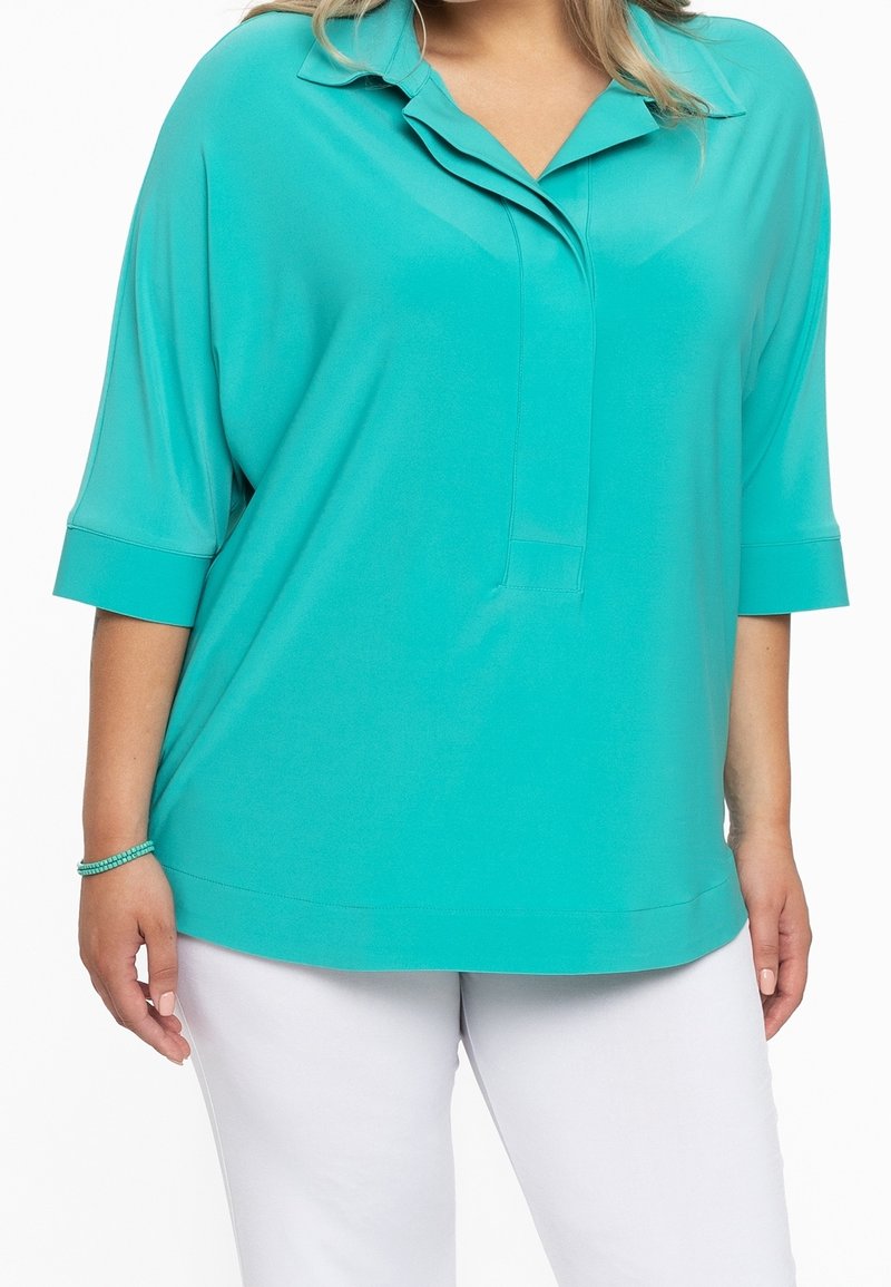 Top turchese, dalla vestibilità ampia, con colletto e maniche corte. Texture morbida, orlo dritto e dettagli delle cuciture discreti. Indossato sopra pantaloni bianchi.