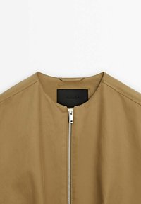 Veste kaki avec un col rond, une fermeture éclair à l'avant et une étiquette en cuir noir. Tissu lisse avec un design minimaliste.