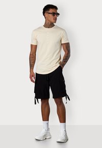 Cremfarbendes Baumwoll-T-Shirt mit vertikalem Text, kombiniert mit schwarzen Cargo-Shorts mit Seitentaschen und Bändern, weißen Sneakers und schwarzen Sonnenbrillen.