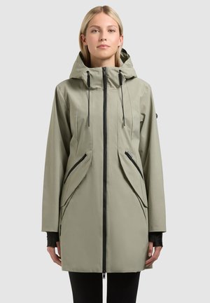 Femeie purtând o haină lungă cu glugă, de culoare verde măsliniu deschis, cu fermoar negru și buzunare cu fermoar oblic, stând pe un fundal simplu.
