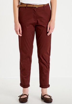 Femme portant un pantalon bordeaux retroussé avec une ceinture tressée marron et des chaussures plates tressées marron, debout sur un fond blanc uni.