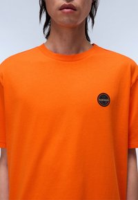 Lys orange bomulds-T-shirt med rund halsudskæring, korte ærmer og et lille sort cirkulært logo i øverste venstre område.