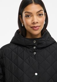 Chaqueta negra acolchada con capucha, presenta botones a presión plateados y un patrón de diamantes en la tela. Textura suave y acentos mínimos.