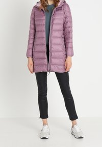 Polstret, lyslilla jakke med hætte, der har lodret syning, lynlås og sidelommer, båret over sorte leggings og sneakers.