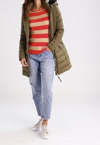 Olijfgroene gewatteerde jas met een bontkraag, rood en beige gestreepte ribbeltrui, versleten lichtblauwe jeans en witte sneakers.