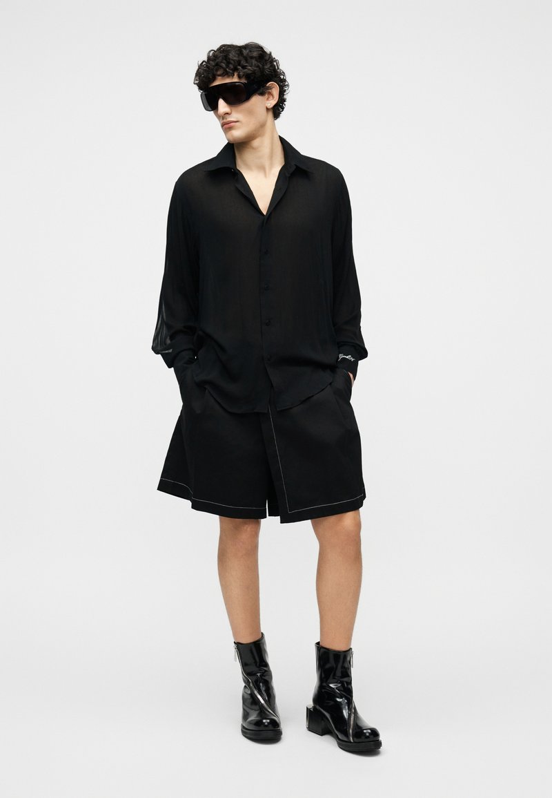 Man wearing oversized black button-up shirt, black knee-length shorts, black patent leather ankle boots, and large black sunglasses.