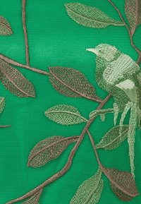 Zelená látka s propracovanou výšivkou listů a ptáka v odstínech hnědé a zelené, s detailními texturami a vzory.