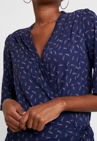 Zbliżenie na osobę ubraną w granatową sukienkę typu wrap w jasnoróżowy wzór kwiatowy oraz duże kolczyki w kształcie obręczy, z dłońmi delikatnie złożonymi.