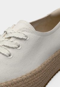 Κοντινό πλάνο ενός λευκού πάνινου αθλητικού παπουτσιού με πάτο από παχύ πλεγμένο σκοινί και λευκά κορδόνια σε μονόχρωμο φόντο.