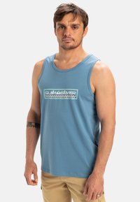 Lyseblå tanktop lavet af bomuld, med et grafisk logo i et ternet mønster i midten. Rund halsudskæring og uden ærmer.