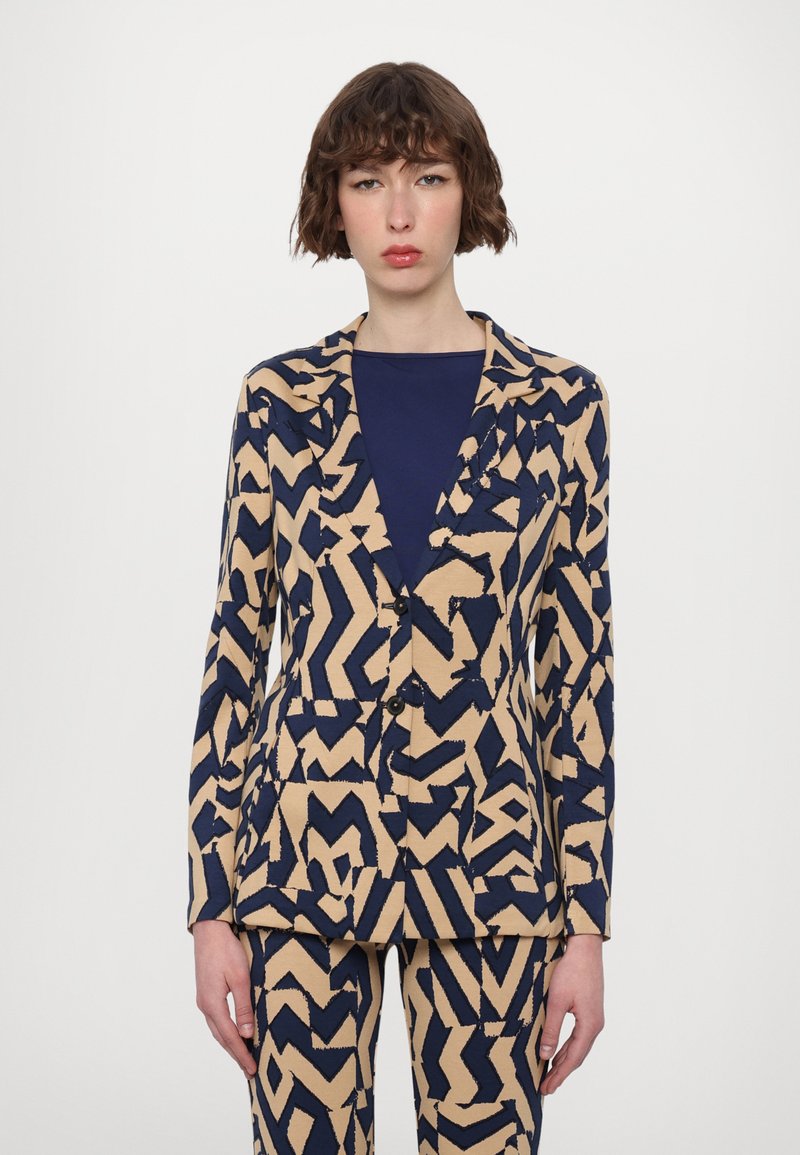 Beige and navy patterned blazer with angular designs, notched lapels, front buttons, and matching trousers. Smooth fabric with a tailored fit.